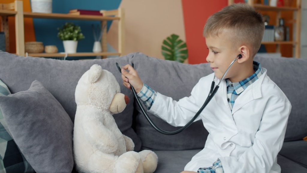 Funny kid is playing with teddy bear examining soft toy with stethoscope enjoying game alone in apartment. Medicine, childhood and house concept.